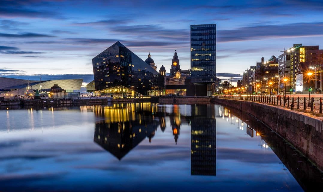 Un moderno lungomare cittadino al crepuscolo, con edifici in vetro illuminati e lampioni che si riflettono nell'acqua calma.