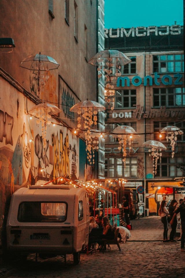 Un vicolo cittadino acciottolato decorato con ombrelli a forma di medusa e luminarie, con un furgone d'epoca parcheggiato di lato.