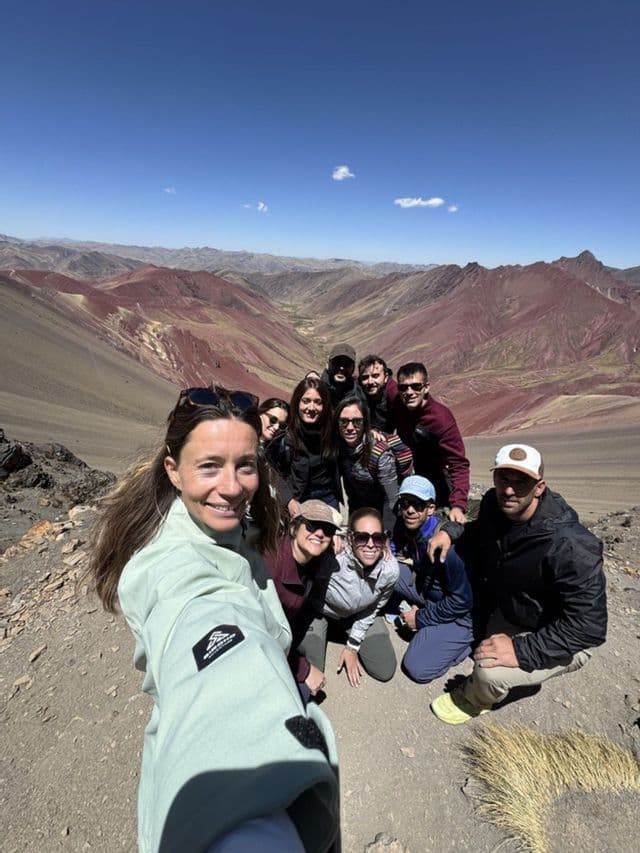 Una mujer se toma una selfie con su grupo de viaje de WeRoad en la cima de una montaña, con vistas a un vasto valle de montañas coloridas y estriadas.