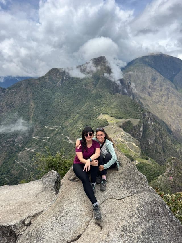 Dos mujeres de un viaje en grupo de WeRoad sonríen mientras están sentadas en una roca con vistas a una vasta cordillera verde bajo un cielo nublado.