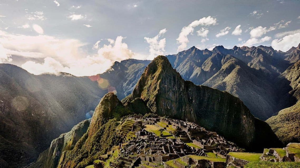 Antiguas ruinas de piedra construidas en una exuberante ladera verde, con altos picos irregulares elevándose al fondo bajo un cielo nublado.