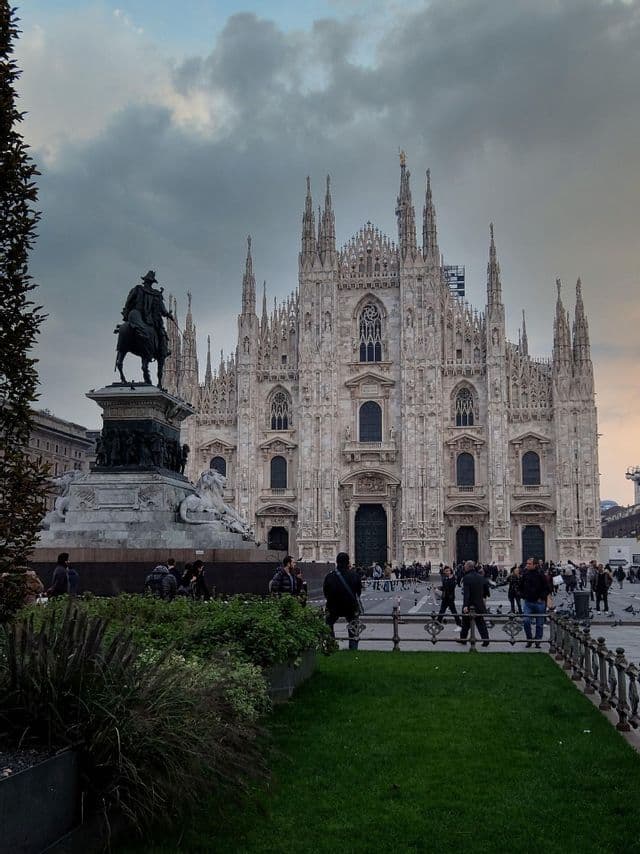 Una ornamentada catedral gótica vista desde una plaza con una estatua ecuestre y gente paseando bajo un cielo nublado.