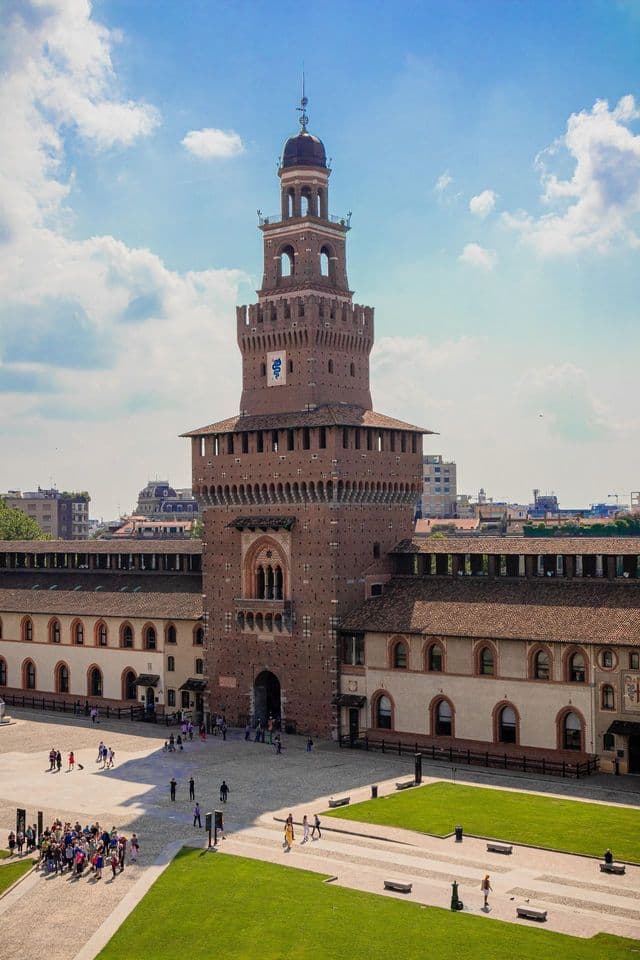Una vista en picada de un gran castillo de ladrillo rojo con una torre central que domina un patio donde la gente camina bajo un cielo parcialmente nublado.