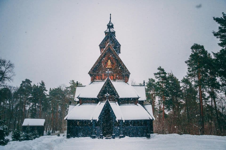 A traditional wooden stave church with snow-covered roofs stands in a forest during a snowfall.