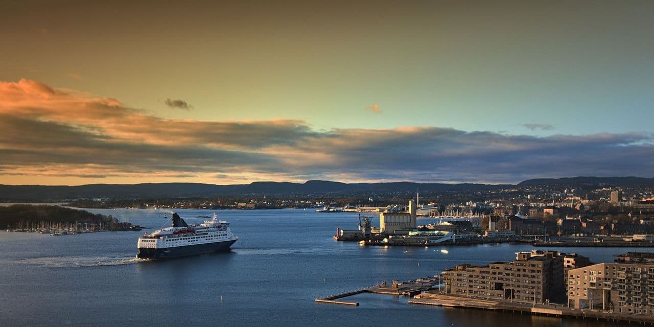 Un grande traghetto naviga in una baia verso una città portuale al tramonto, con colline lontane sotto un cielo nuvoloso.