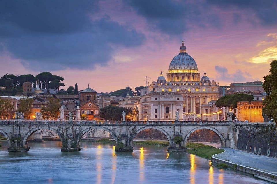 Un ponte in pietra ad arco attraversa un fiume, conducendo a una grande basilica illuminata al tramonto.