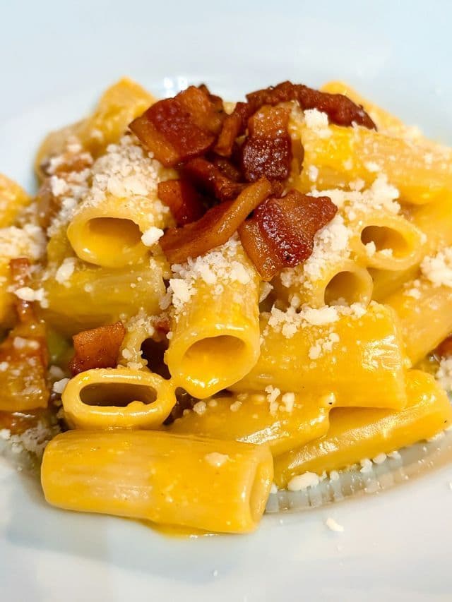 Primo piano di rigatoni in salsa cremosa gialla, conditi con pancetta croccante e formaggio grattugiato.