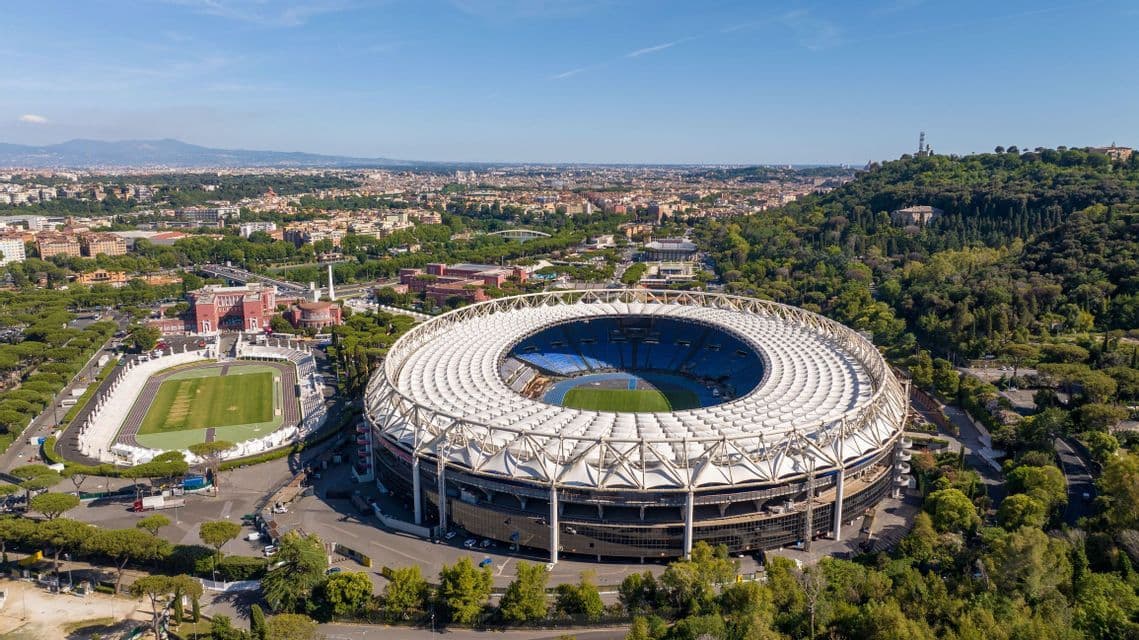 Una vista aerea di un grande stadio circolare accanto a una pista di atletica, con una città e delle colline visibili sullo sfondo.