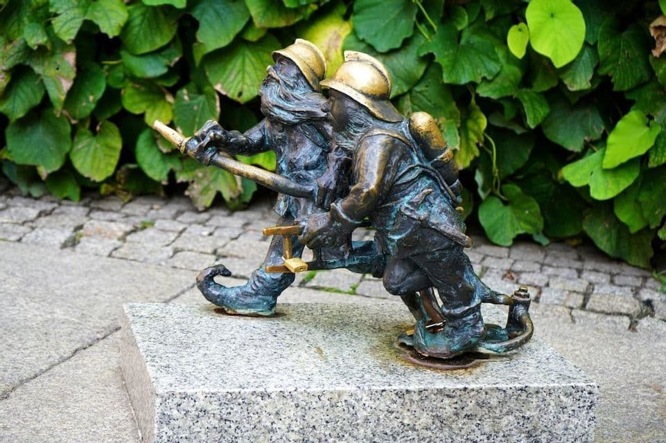 Una piccola statua in bronzo raffigurante due gnomi vestiti da pompieri che reggono una manichetta antincendio, su un piedistallo di pietra con uno sfondo di foglie verdi.