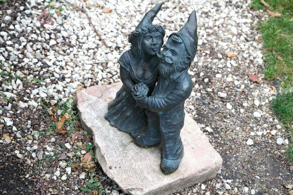 Una scultura in metallo scuro di una coppia di gnomi si erge su una pietra in un giardino di ghiaia, con il maschio che tiene una rosa.