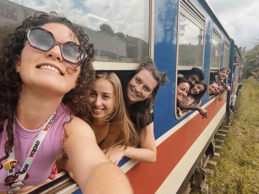 Un groupe WeRoad prend un selfie en souriant et en se penchant par les fenêtres d'un train bleu et rouge.