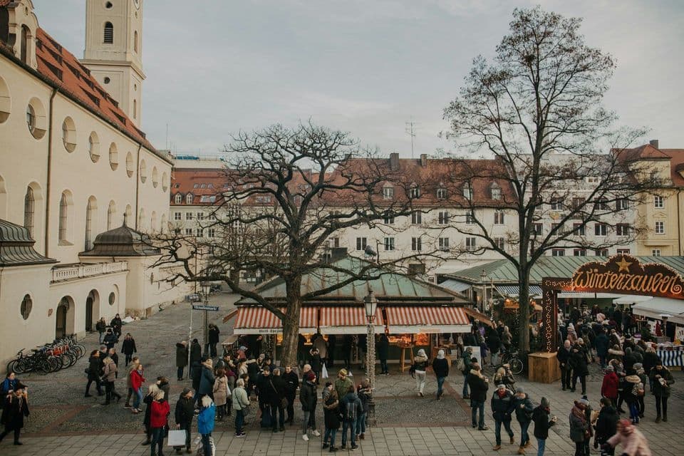 Vista aerea di un vivace mercato invernale in una piazza acciottolata, con persone che curiosano tra le bancarelle accanto a edifici storici.