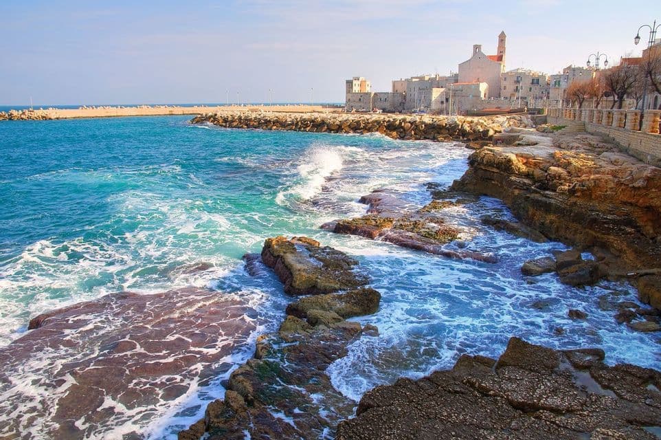 Turquoise waves crash against a rocky coastline in front of a historic stone town under a clear blue sky.