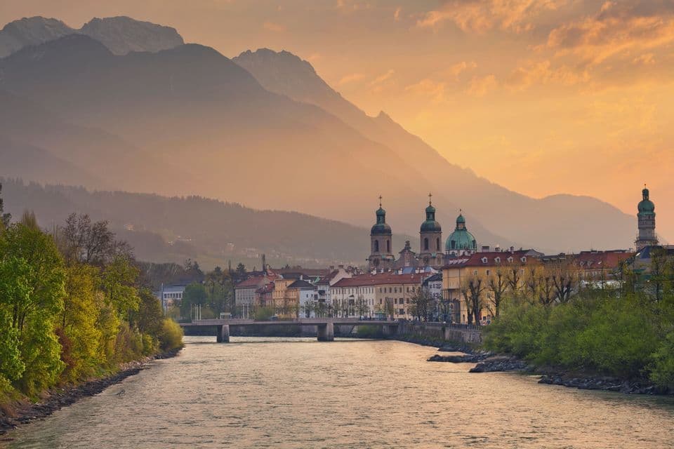 Eine Stadtansicht mit Kuppeltürmen liegt an einem Flussufer mit einer Brücke, im Hintergrund neblige Berge unter einem goldenen Himmel.