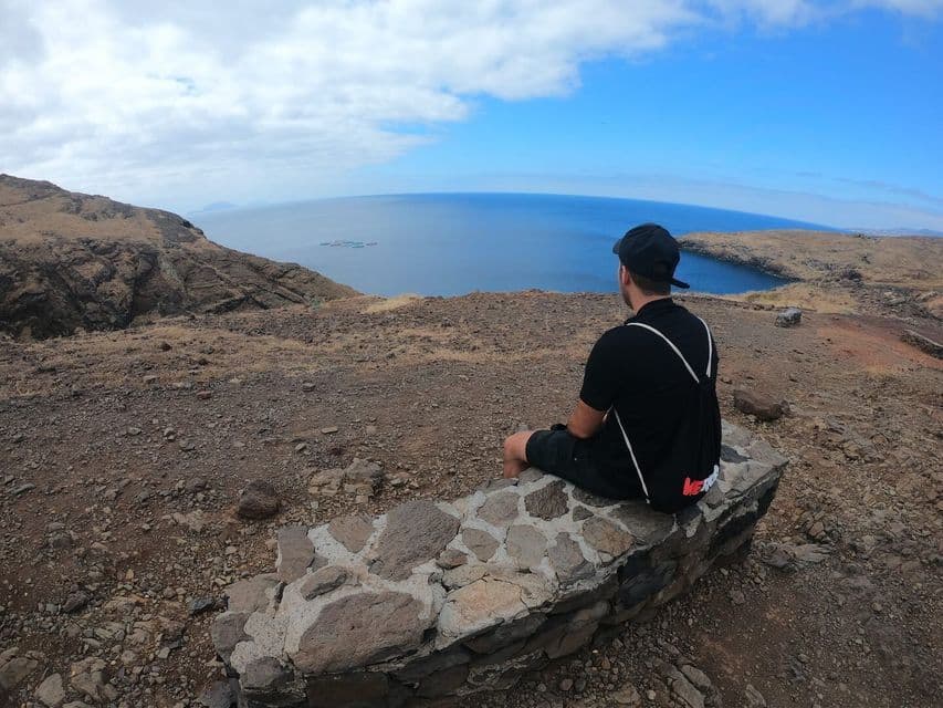 Un hombre visto de espaldas se sienta en un muro bajo de piedra en un acantilado, mirando el océano en un día parcialmente nublado.