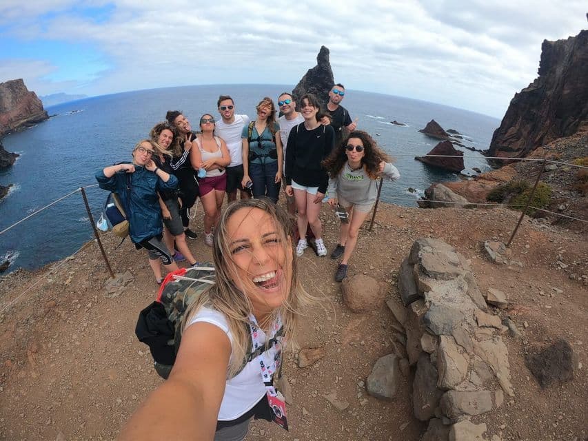 Un groupe WeRoad prend un selfie grand-angle sur une falaise surplombant l'océan et des formations rocheuses.