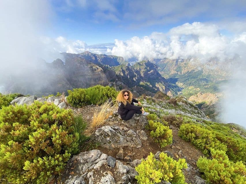 A woman with curly hair sits on a rocky peak overlooking a vast mountain valley partially covered by clouds.