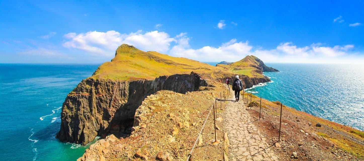 Ein Wanderer mit Rucksack wandert auf einem schmalen Steinpfad entlang einer hohen, grasbewachsenen Küstenklippe mit Blick auf das Meer.