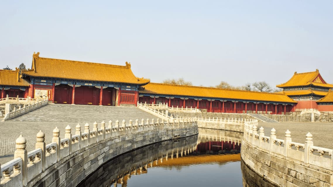 Una balaustra in pietra curva corre lungo un fossato, riflettendo edifici tradizionali cinesi con tetti giallo-oro.