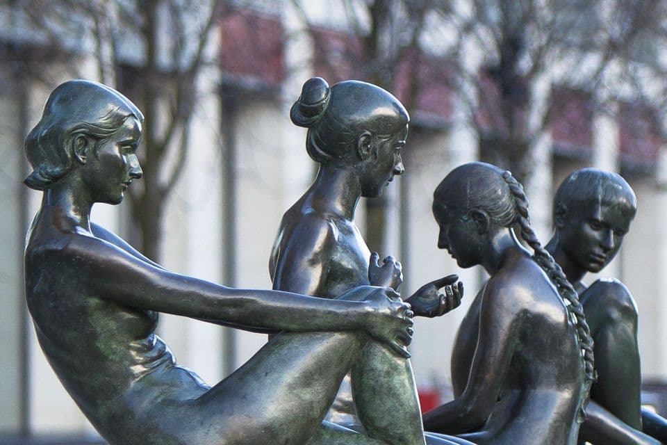 Un primer plano detallado de una escultura de bronce oscuro que representa a cuatro figuras femeninas sentadas juntas en un entorno al aire libre.