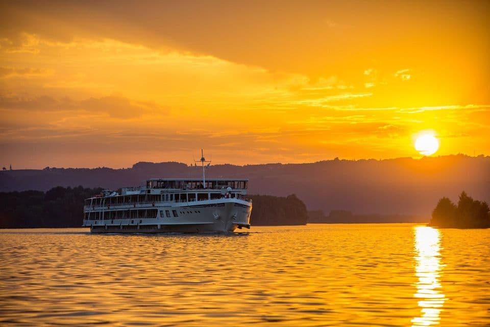 Un crucero blanco navega en aguas tranquilas durante una puesta de sol dorada, con colinas silueteadas al fondo.