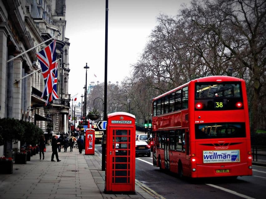Un autobús rojo de dos pisos circula por una calle de la ciudad junto a una cabina telefónica roja, con una bandera Union Jack exhibida en un edificio.