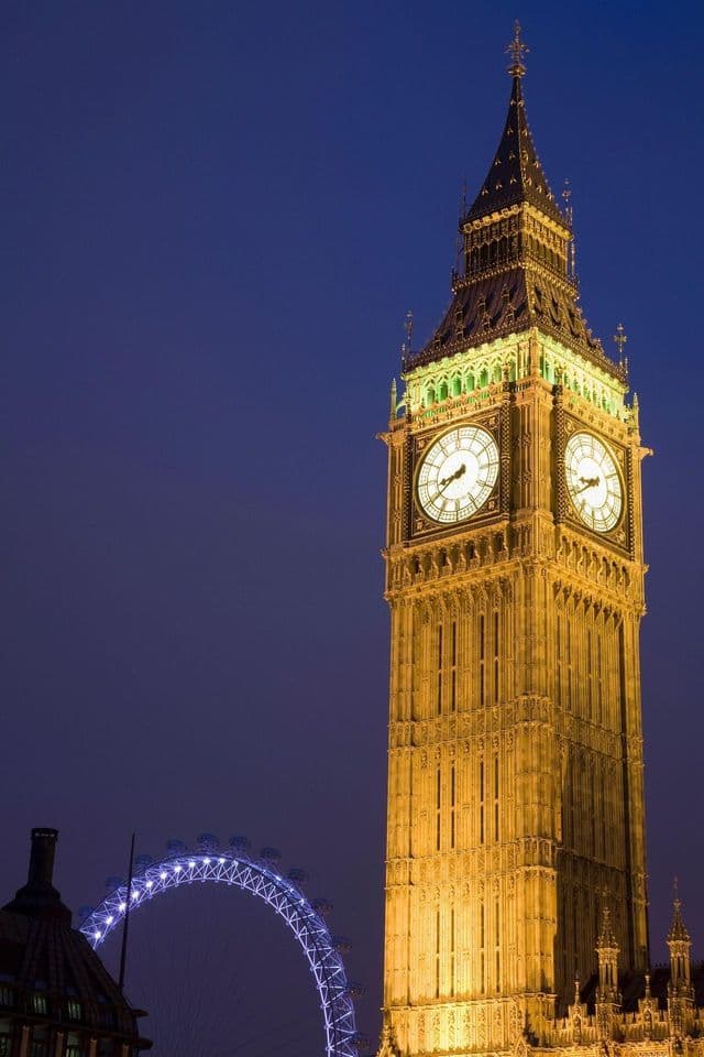 La iluminada torre del reloj Big Ben se alza contra un cielo nocturno azul oscuro, con el London Eye visible al fondo.