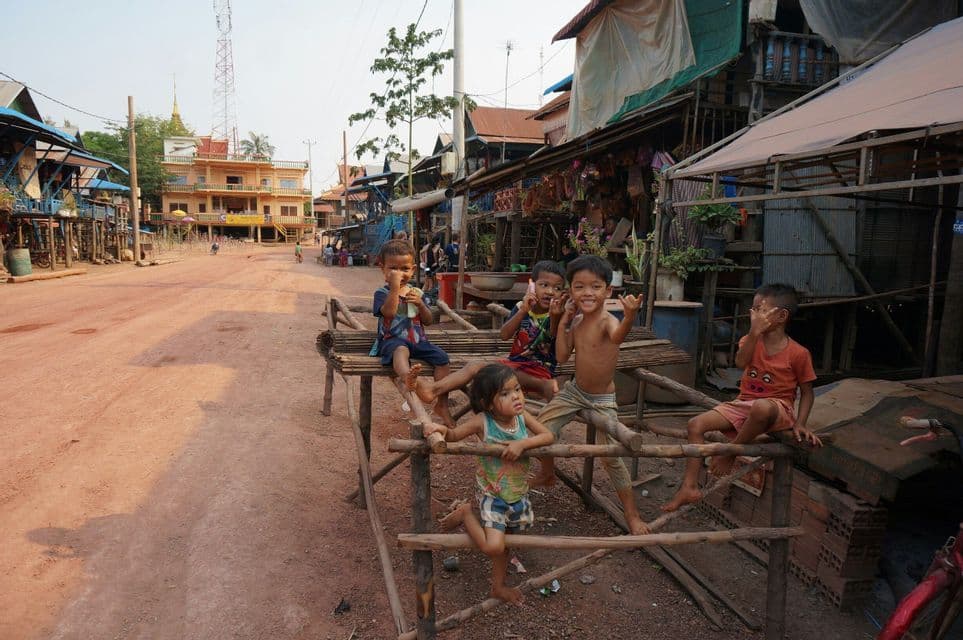 Un gruppo di bambini sorride e posa per la macchina fotografica, seduti su una struttura di legno lungo una strada sterrata in un villaggio.