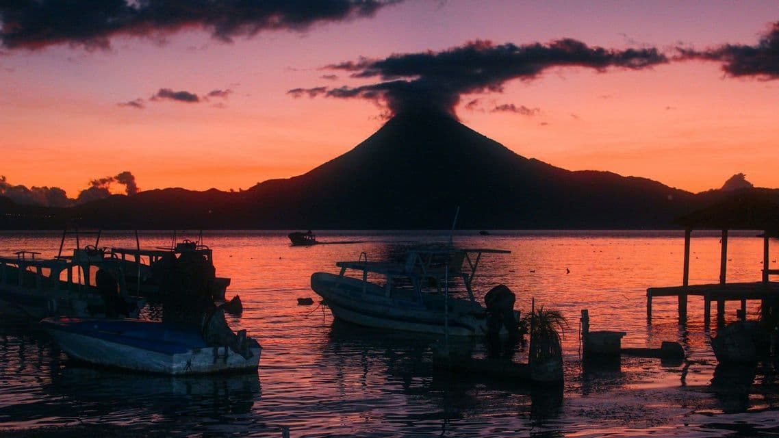 Un vulcano in silhouette si staglia su un lago con barche che galleggiano sull'acqua al tramonto colorato.