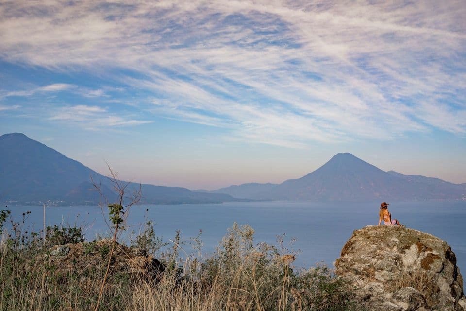 Una persona con un cappello siede su una grande roccia che domina un vasto lago e montagne distanti sotto un cielo azzurro con nuvole leggere.