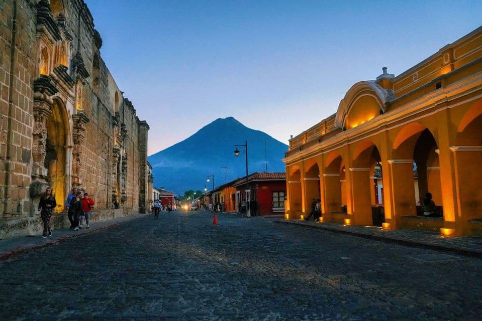Una strada acciottolata affiancata da edifici coloniali illuminati conduce verso un grande vulcano al tramonto.