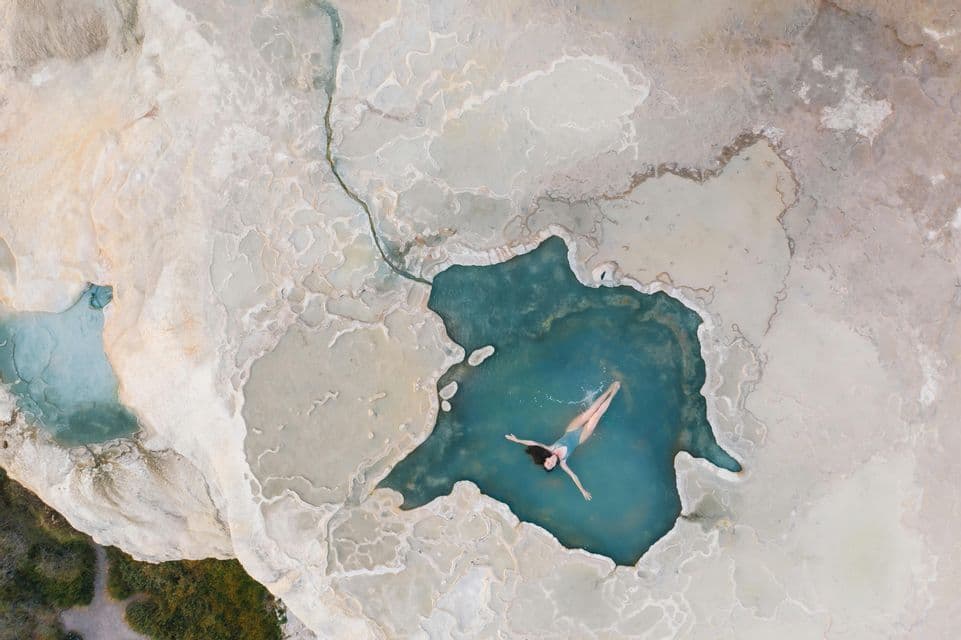Vista aerea dall'alto di una donna che galleggia sulla schiena in una piscina naturale turchese circondata da terrazze minerali bianche.