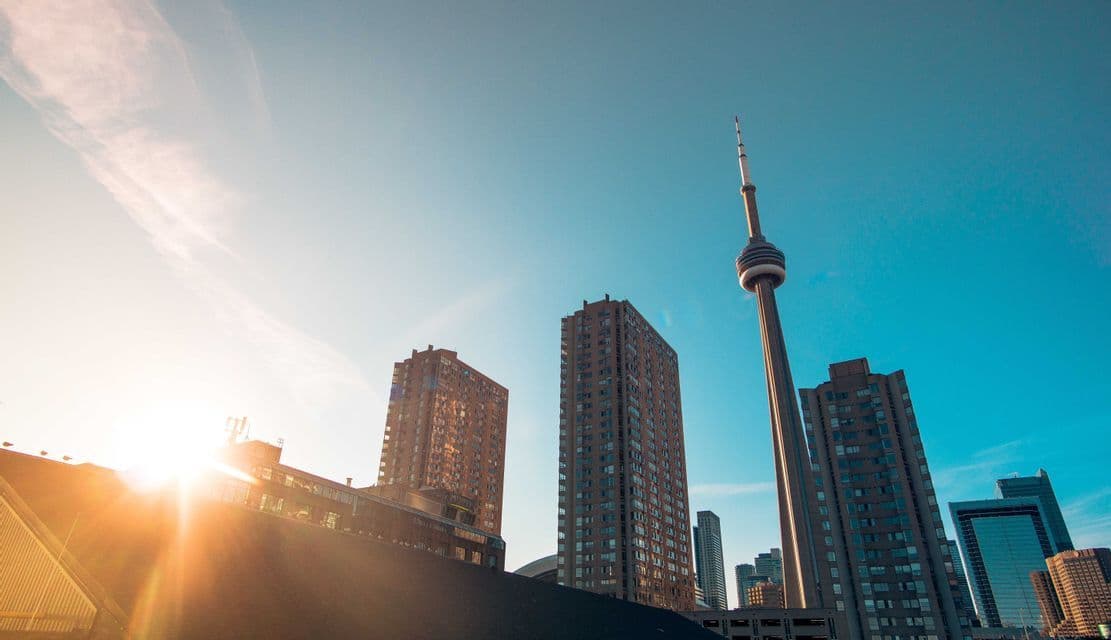 A low-angle shot of a city skyline with a prominent observation tower and several high-rise buildings against a clear blue sky with a sun flare.