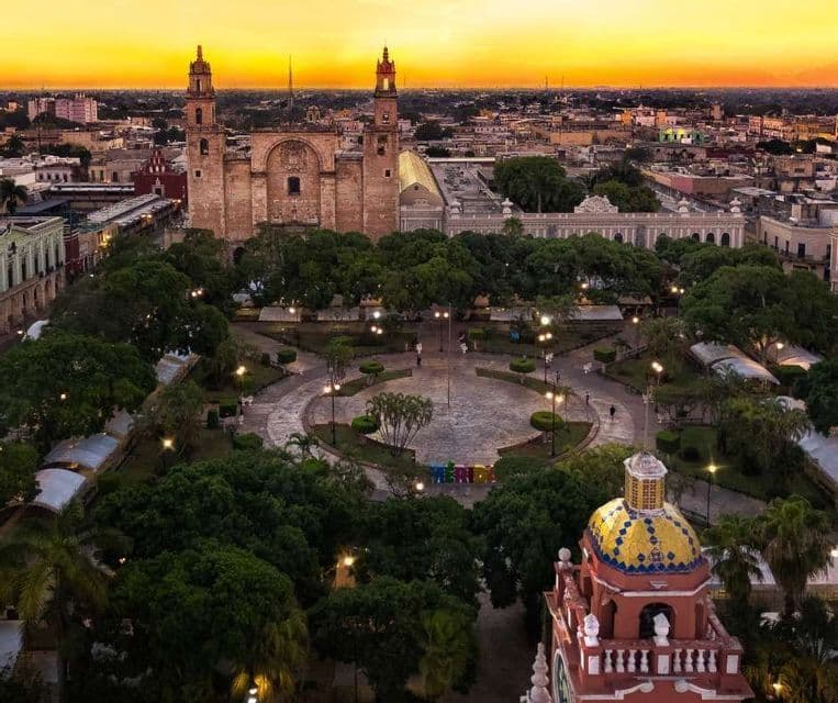 An aerial view of a historic cathedral with two bell towers overlooking a city plaza filled with trees at sunset.