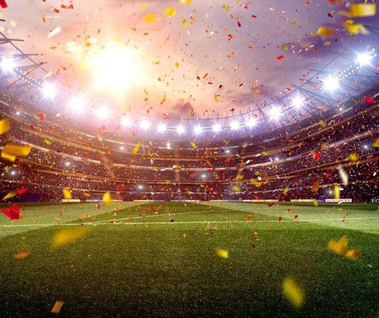 Colorful confetti falls onto the green grass field of a packed sports stadium illuminated by bright floodlights.