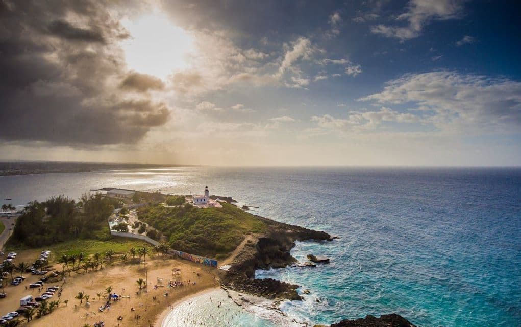 Una vista aérea de un faro en un promontorio verde dominando una playa de arena y el océano azul bajo un cielo parcialmente nublado.