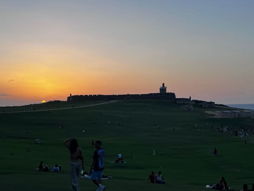 Personas relajándose en una gran colina de hierba, viendo el atardecer tras una fortaleza costera histórica con un faro.
