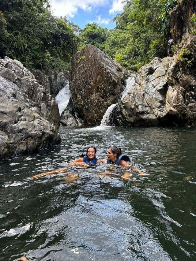Dos mujeres de un viaje en grupo de WeRoad sonriendo mientras nadan en una poza natural al pie de una cascada rocosa.