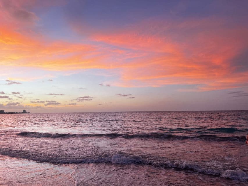 El océano refleja un atardecer colorido mientras suaves olas rompen en la orilla, con un horizonte urbano visible en la distancia.