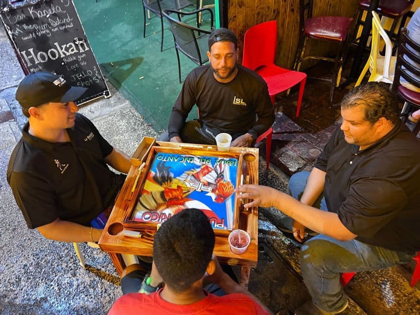 Cuatro hombres se sientan alrededor de una mesa de madera jugando una partida de dominó por la noche en un tablero colorido con temática puertorriqueña.