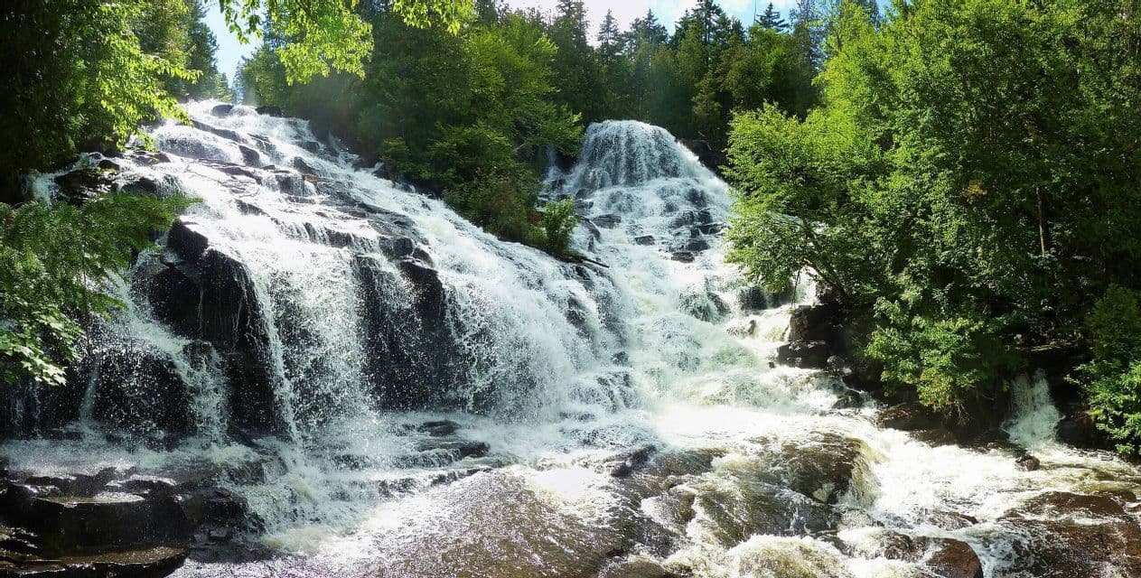 A wide, multi-tiered waterfall cascades down a rocky slope, surrounded by a dense green forest under a bright sky.
