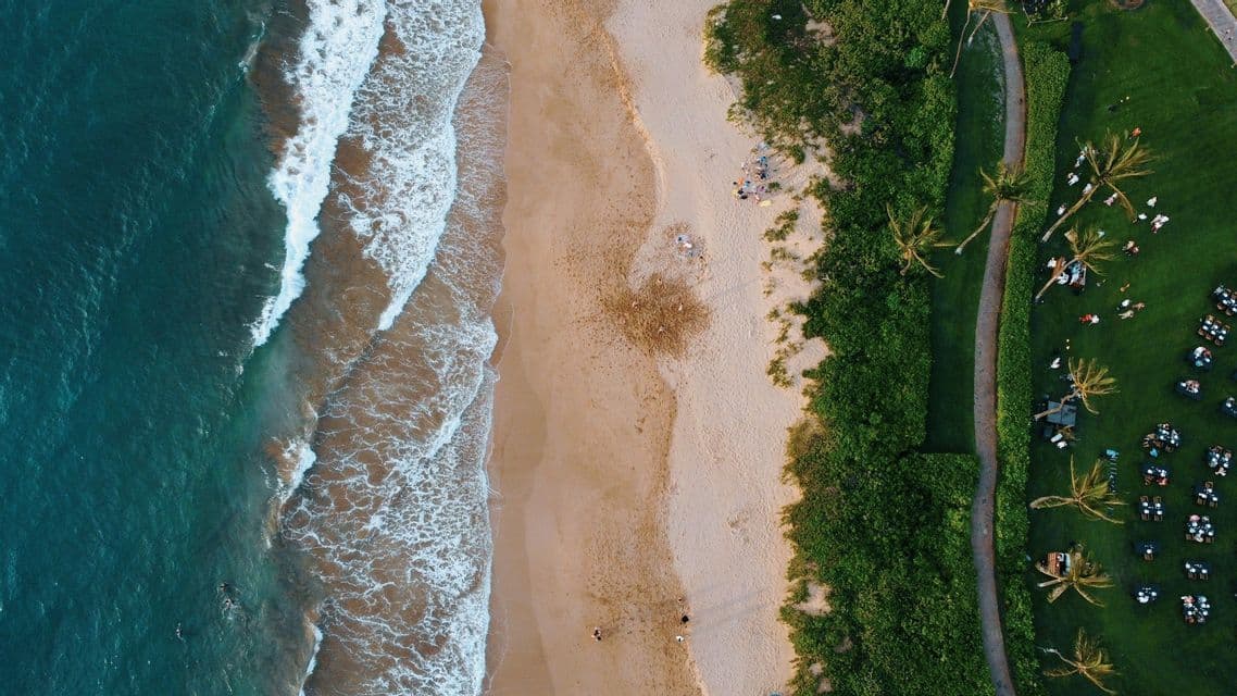 Vista aérea cenital de una costa, con olas turquesas del océano, una playa de arena y un césped verde con mesas preparadas para un evento.