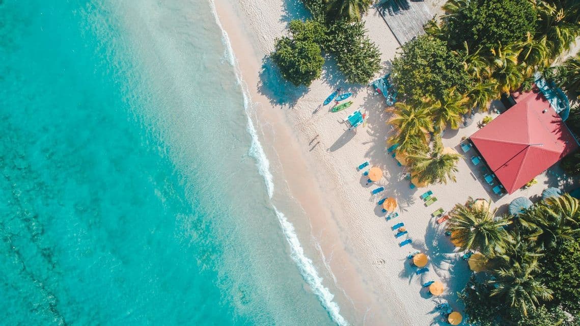Vista aérea cenital de una playa tropical con agua turquesa, arena blanca, árboles frondosos y un edificio con techo rojo.