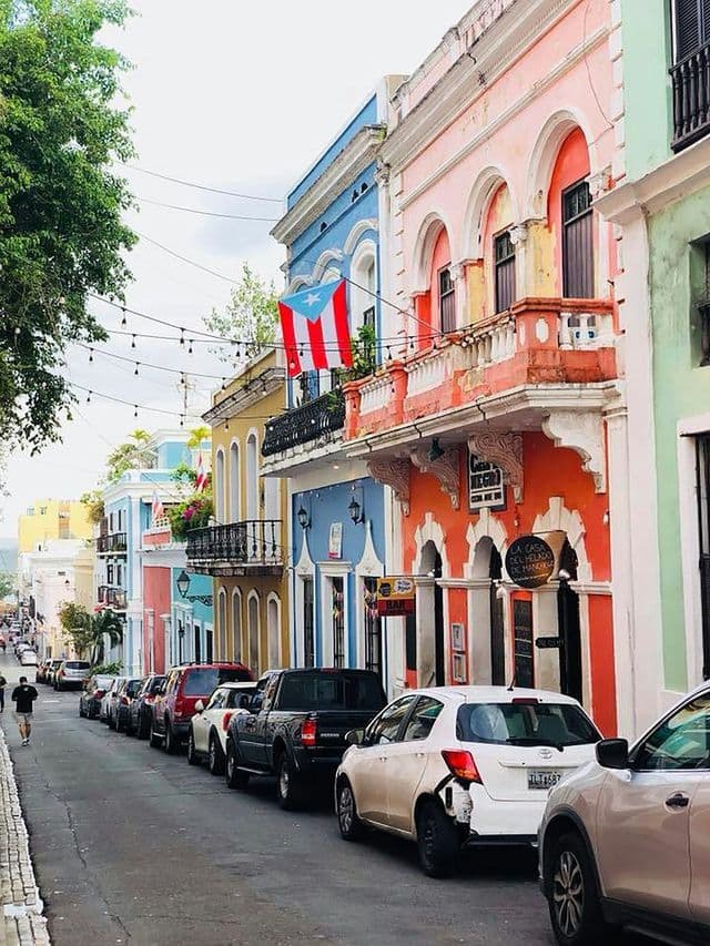 Una bandera puertorriqueña cuelga de un balcón en una calle estrecha llena de edificios coloridos y coches aparcados.