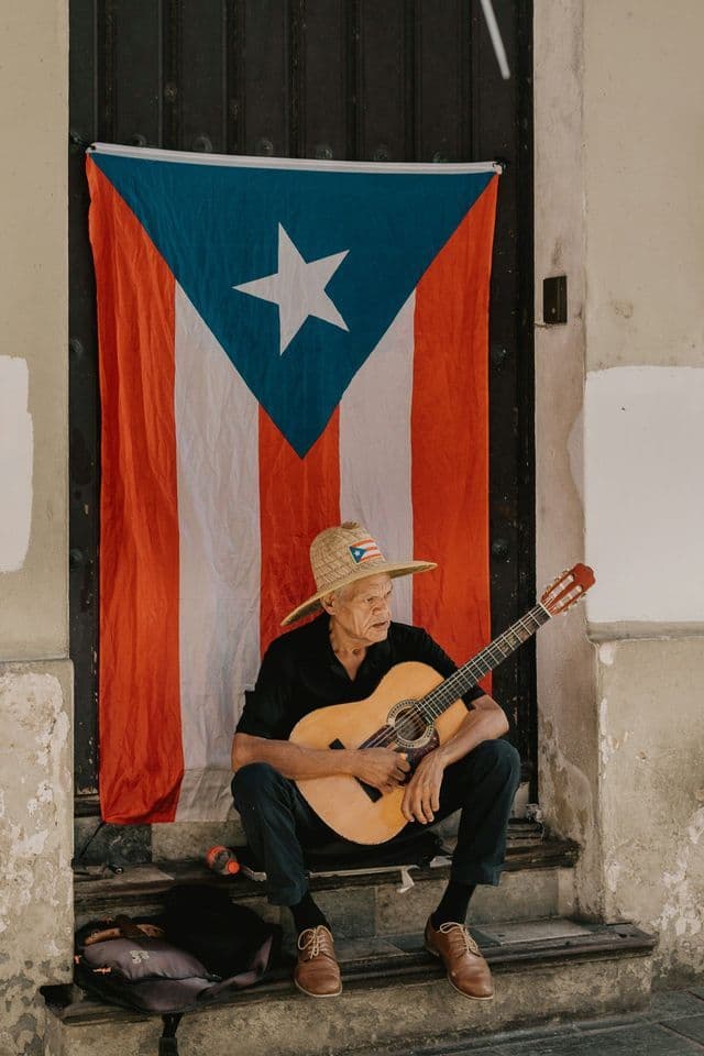 Un anciano con un sombrero de paja toca una guitarra clásica mientras está sentado en unas escaleras de piedra frente a una gran bandera puertorriqueña.