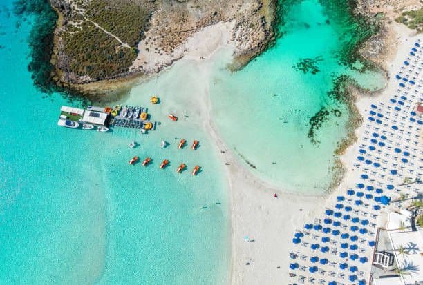 Vue aérienne d'une crique de sable avec une eau turquoise limpide, des rangées de parasols bleus et une jetée flottante avec des bateaux.