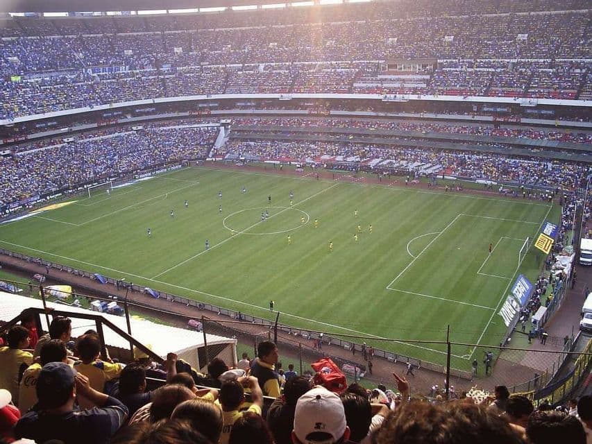 A high-angle view of a football match being played in a large, multi-tiered stadium packed with spectators under bright sunlight.
