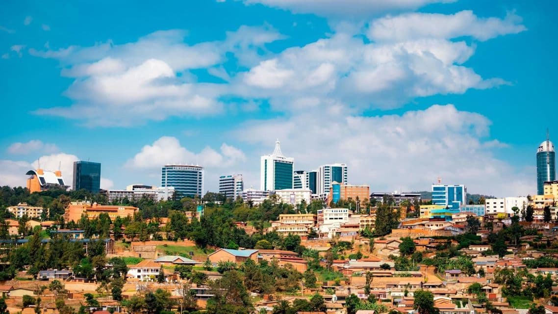 Un panorama urbain de gratte-ciel modernes et d'immeubles de bureaux s'élevant derrière des maisons traditionnelles sur un flanc de colline verdoyant sous un ciel partiellement nuageux.