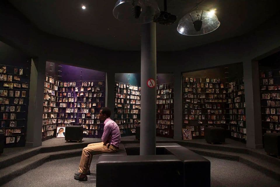 Un homme est assis sur un banc dans une salle commémorative circulaire faiblement éclairée, contemplant les murs recouverts de rangées de portraits photographiques.