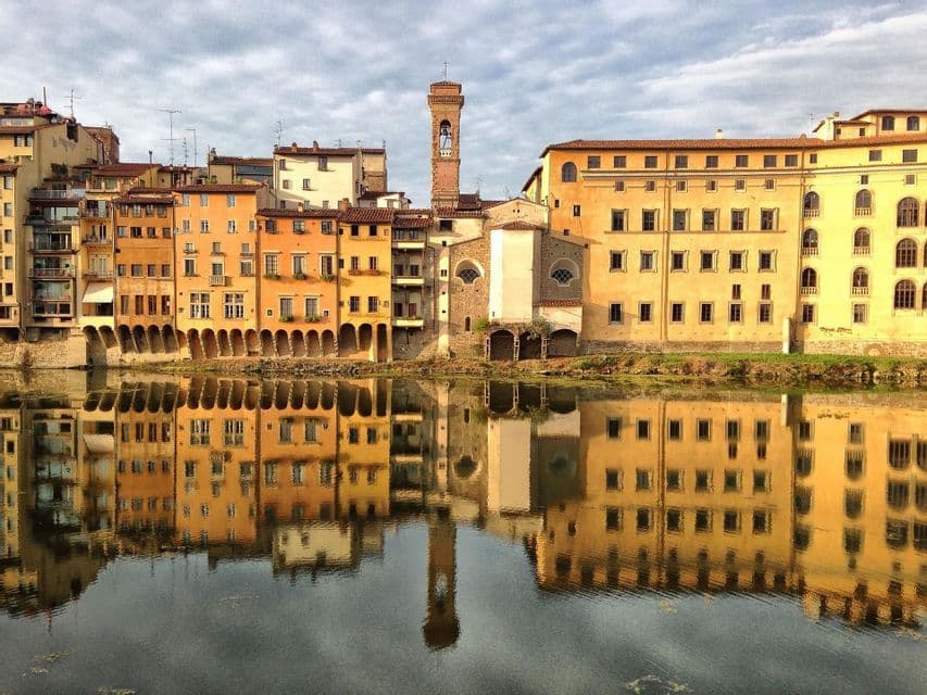 Coloridos edificios históricos y un campanario a lo largo de un río, perfectamente reflejados en el agua tranquila bajo un cielo nublado.